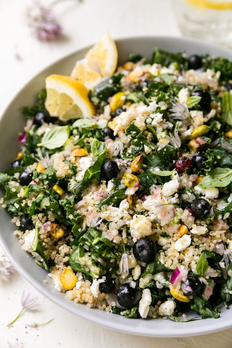 Quinoa kale salad with blueberries and feta in white bowl with lemon wedge on side.