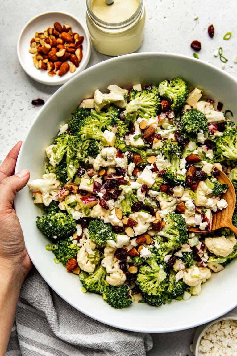 Large serving bowl filled with apple broccoli cauliflower salad tossed in lemony dressing being held by two hands.